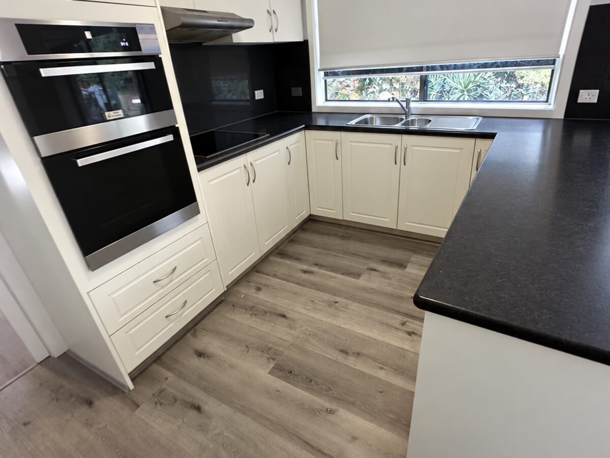 Dark Grey Oak 1005 planks in a kitchen with cream shaker cabinetry, a black stone benchtop and stainless wall ovens.