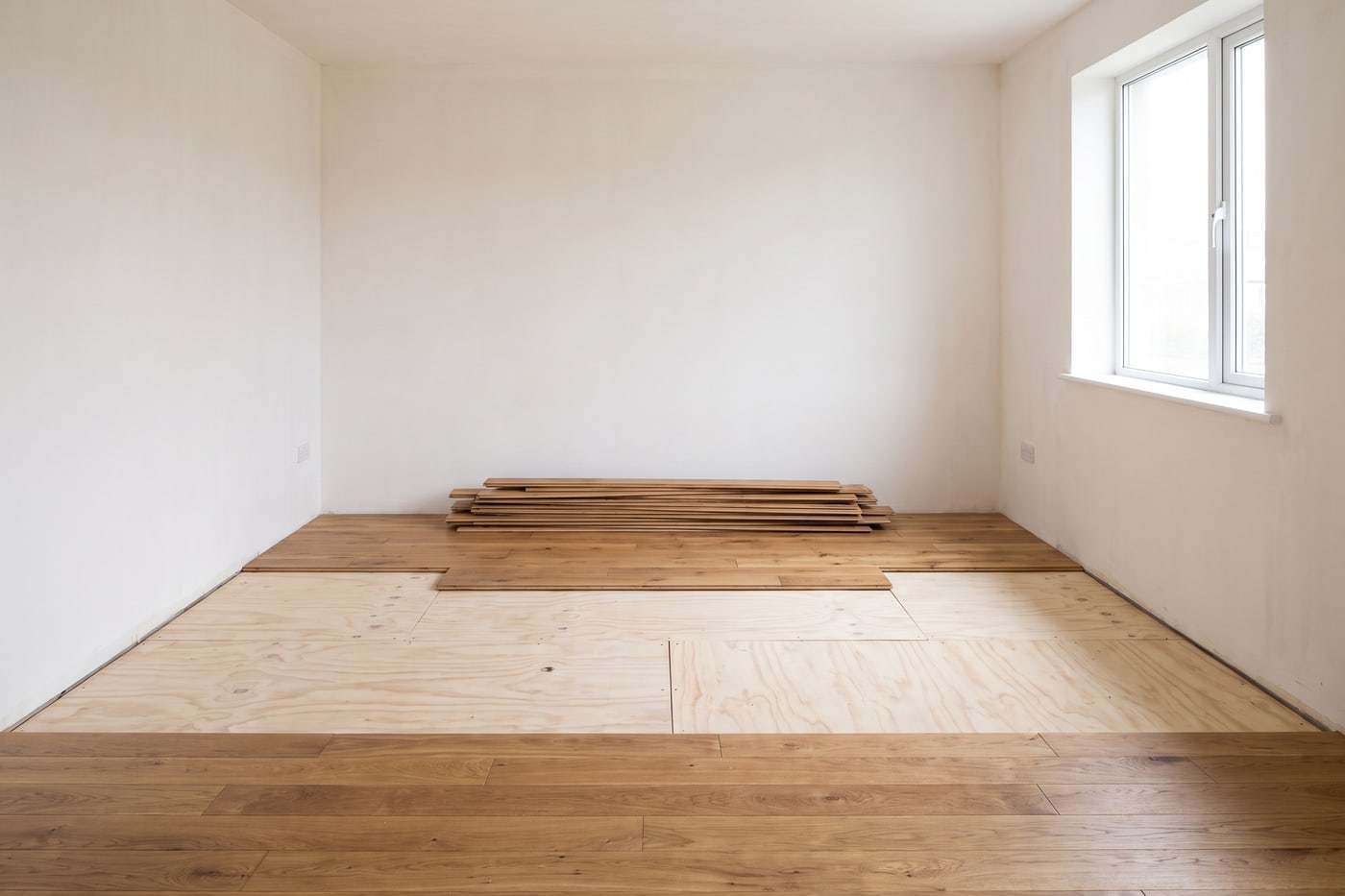 An empty room mid-renovation, a section of floating floor lifted to reveal plywood subfloor, boards stacked to one side