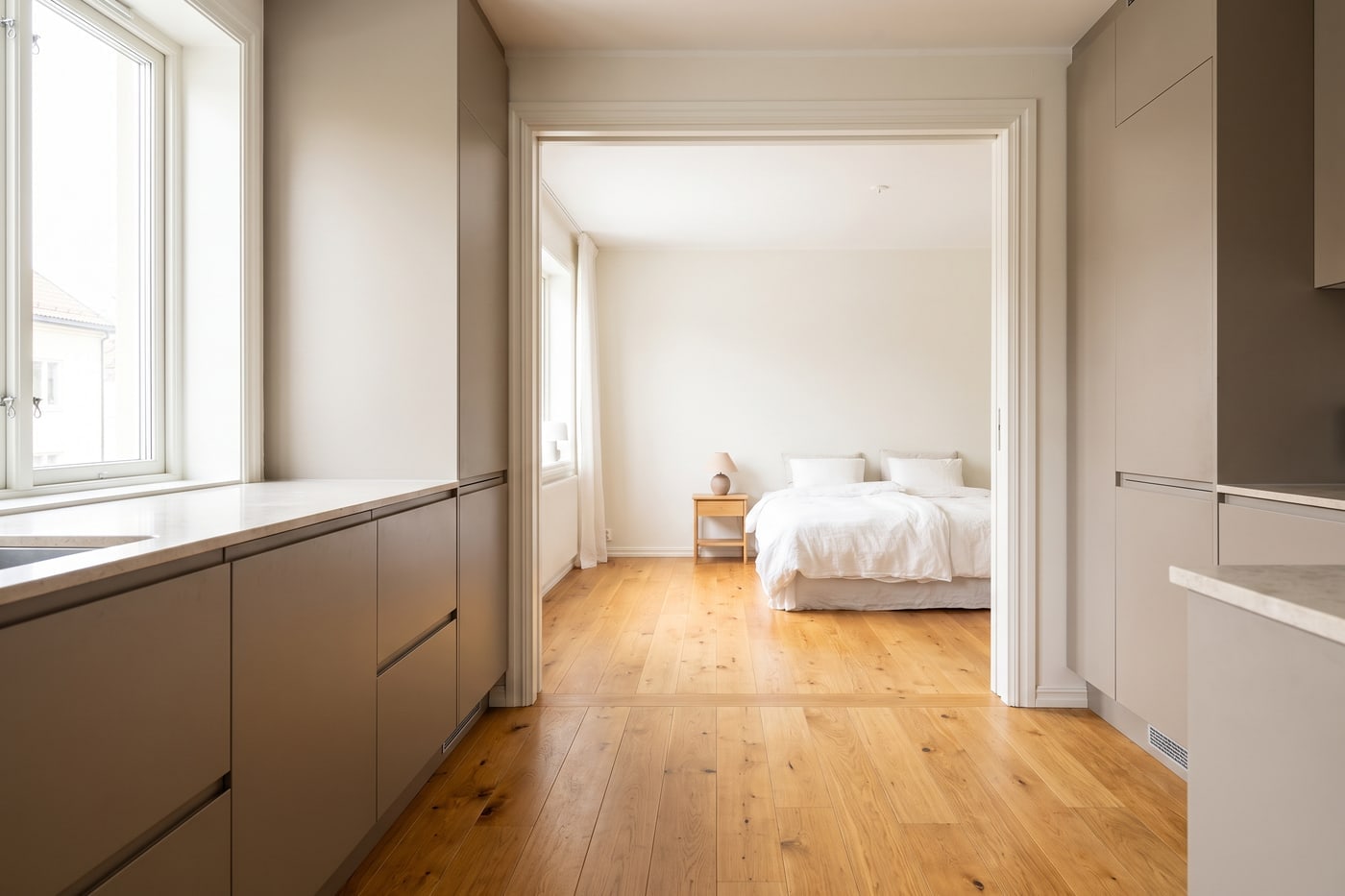 A kitchen and a bedroom seen through a wide doorway, with the same timber-look flooring running continuously through both